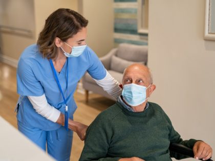 Friendly nurse wearing protective face mask helping handicapped senior man in wheelchair a